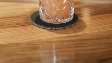 A refreshing cocktail in an elegant glass with a slice of lemon and mint. The drink is placed on a wooden counter, with a blurred bar visible in the background.