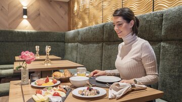 A woman is sitting at a table in an elegant restaurant and enjoying a hearty breakfast. On the table, various fruits, pastries, and a drink are arranged.