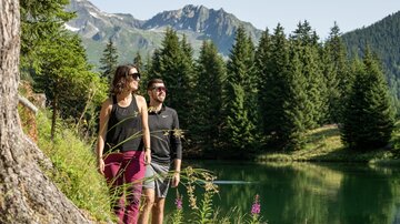 A couple is hiking along the shore of a clear lake, surrounded by tall trees and mountains. The atmosphere is sunny and inviting.