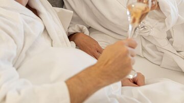 A couple in bathrobes sits together and toasts with glasses of champagne. They smile and enjoy a relaxing moment.