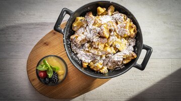 A tasty dish made of small, golden pieces, dusted with powdered sugar, served in a black pot. Accompanied by a small bowl of applesauce and fresh berries.