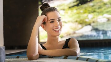 A young woman smiles relaxingly in the water. In the background, green trees can be seen.
