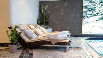A relaxing lounge bed with pillows is next to a pool. In the background, plants and a bright wall can be seen.