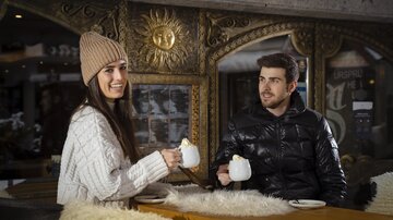 A couple is sitting in a cozy environment enjoying hot drinks. The woman is wearing a warm sweater and hat, while the man is smiling in a jacket.