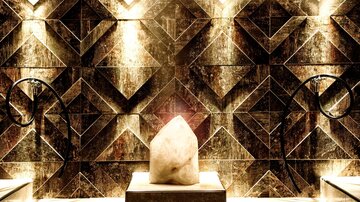 An elegant room with geometric mosaic patterns on the wall. In the center stands a large, bright stone illuminated by soft lights.