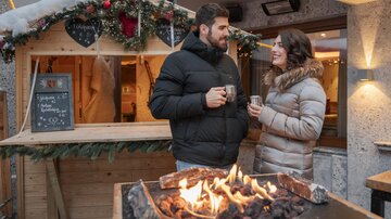 A couple stands in front of a festively decorated booth during the winter season. They are holding cups in their hands and smiling at each other.