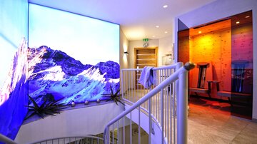 A modern wellness lounge with a large, colorful screen displaying a mountain landscape. In the background, sauna and relaxation areas can be seen.