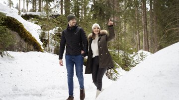 A couple walks through a snowy landscape in the forest. The woman points up with her hand while the man walks beside her.