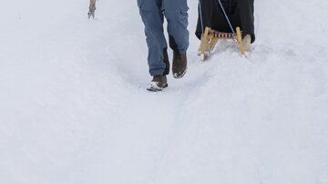 A man pulls a sled through a snowy landscape, while a woman joyfully sits on the sled. In the background, snow-covered mountains and trees can be seen.