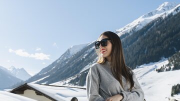 A woman in sunglasses stands by a railing and smiles. In the background, snow-covered mountains stretch out under a clear blue sky.