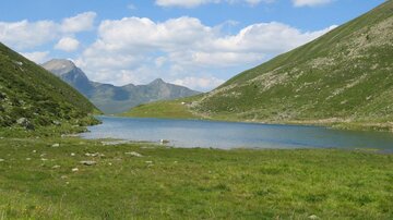 A quiet lake, surrounded by green meadows and gentle hills. In the background, mountains and a blue sky can be seen.