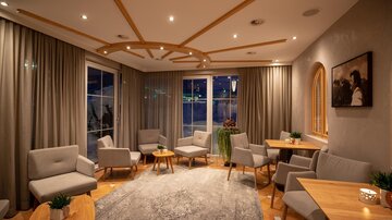 A modern living room with comfortable armchairs and wooden tables. Large windows offer a view outside, while soft light creates a welcoming atmosphere.