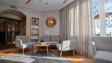 A bright and inviting lounge with modern furniture and a large window. Soft curtains and a tiled floor create a cozy atmosphere.
