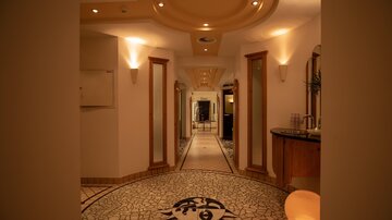 An elegant hallway with a decorative floor mosaic and warm light. The walls are bright, and there are several doors along the corridor.