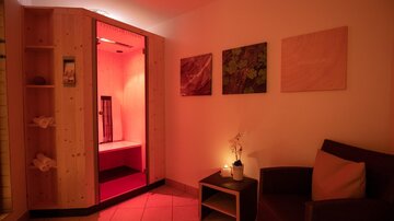 A cozy room with a sauna and soft red lighting. On the left, towels are ready, and a comfortable armchair invites you to relax.