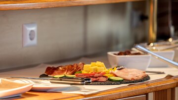 A rich breakfast buffet with various types of cheese, cold cuts, and fresh fruit. The selection is displayed on a dark plate in a warm setting.