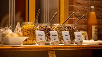 A buffet with various jars full of cereals and muesli. Next to it is a bowl and a bottle of fruit juice.