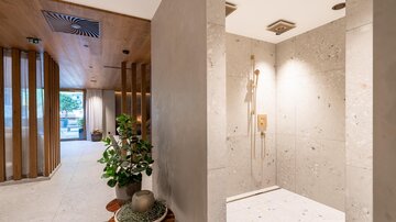 A modern bathroom with a stone shower and elegant fixtures. In the background, plants and a brightly lit room are visible.