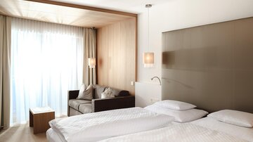 A modern hotel room with two beds and a cozy seating area. Large windows provide plenty of daylight.