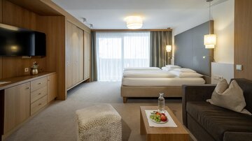A modern hotel room with two single beds, a television, and a comfortable sofa. Large windows provide plenty of natural light.