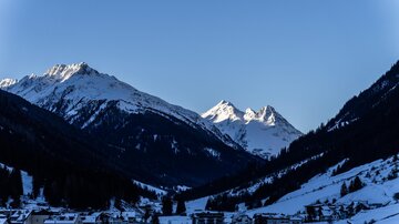 A tranquil winter landscape with snow-covered mountains and a clear blue sky. In the foreground, small houses are visible in a valley.