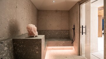 A modern steam room with elegant stone walls and soft lighting. A pink quartz sculpture decorates the seating area.