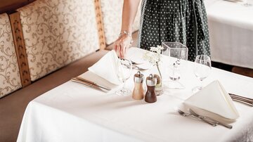 An elegantly set table in a restaurant. A person brings dishes and glassware to the table.