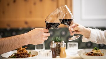 Two people toast with red wine glasses. On the table are two plates with delicious food.