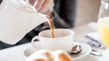 A hand pours freshly brewed tea into a white cup. In the foreground, a roll is resting on a plate.