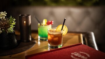Two cocktails on a wooden table in a cozy environment. In the foreground, a drink with an orange and in the background, a green cocktail.