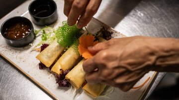 A plate with spring rolls is being decorated. There are also two dips and fresh vegetables.