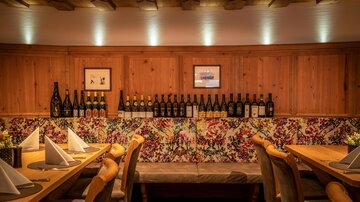 A cozy restaurant with wooden walls and colorful flower patterns. On a shelf, many bottles are arranged, and the tables are tastefully set.